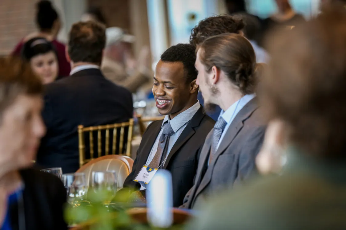 Out of focus people sit at tables attending an event. A young man is in focus in the center of the photo. He has dark brown skin and is wearing a suit. He is smiling and speaking with another student wearing a suit. He looks happy and proud. .