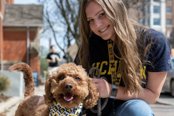 A light skinned girl in a Michigan shirt crouches down next to a brown labradoodle wearing a Michigan bandana.