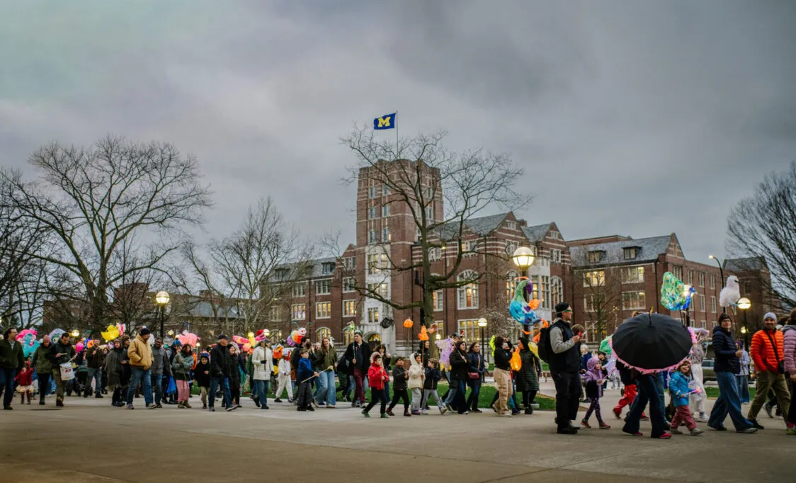 It’s a cloudy evening. In the foreground a parade of children holding bright lanterns are walking by. The Michigan Union is in the background.
