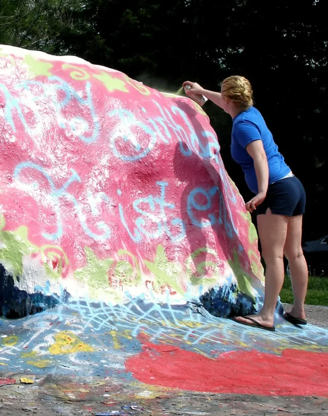 The Rock, an off campus landmark, is a large rock that students spray paint. A student in a blue shirt is paining the rock pink