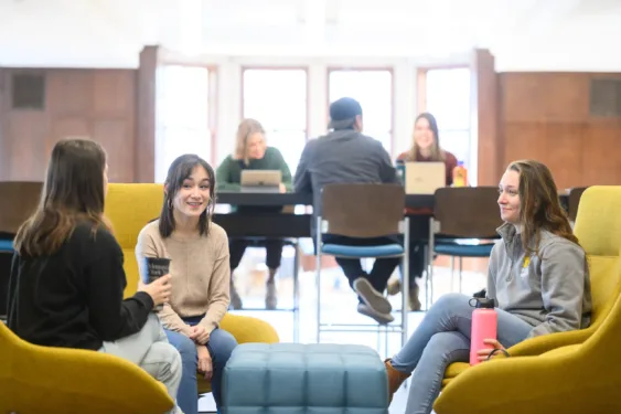 Three students sitting in lounge chairs at the Michigan Union talking and laughing. In the background a group of students work at a table and light filters through the windows.