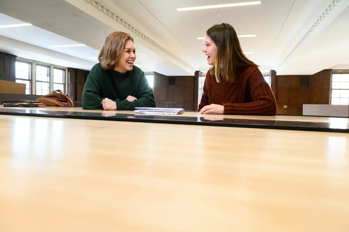 Two students talking and smiling at a light oak table with sunlight filtering in from the left.