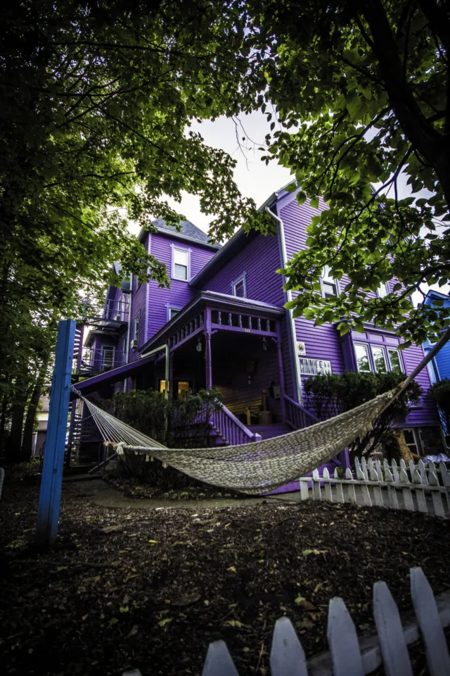 A low-angle shot of a large, multi-story Victorian-style house painted entirely in vibrant purple, with a woven rope hammock strung across the front yard under leafy green trees.