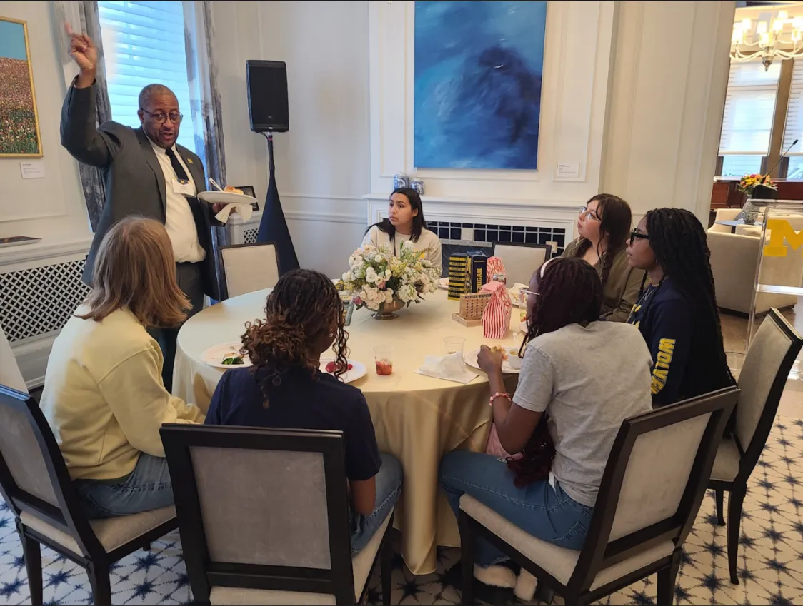 Vice president for student life Martino Harmon speaks to a group of students seated around a circular table. His arm is raised in the air as he talks.