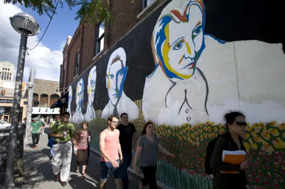 A group of people walk past a large wall mural on Liberty Street. The mural depicts the headshots of Woody Allen, Edgar Allan Poe, Hermann Hesse, Franz Kafka, and Anaïs Nin.