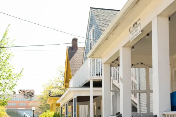 The front porches of three off-campus housing units. Trees and bushes line the front yards and the sun shines brightly overhead.