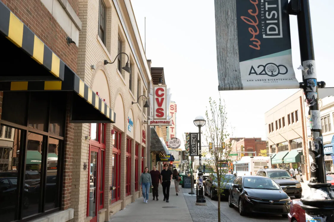 People walking down the sidewalk at State Street, with the State theater in the background. There are signs for various businesses and cars pack the street.