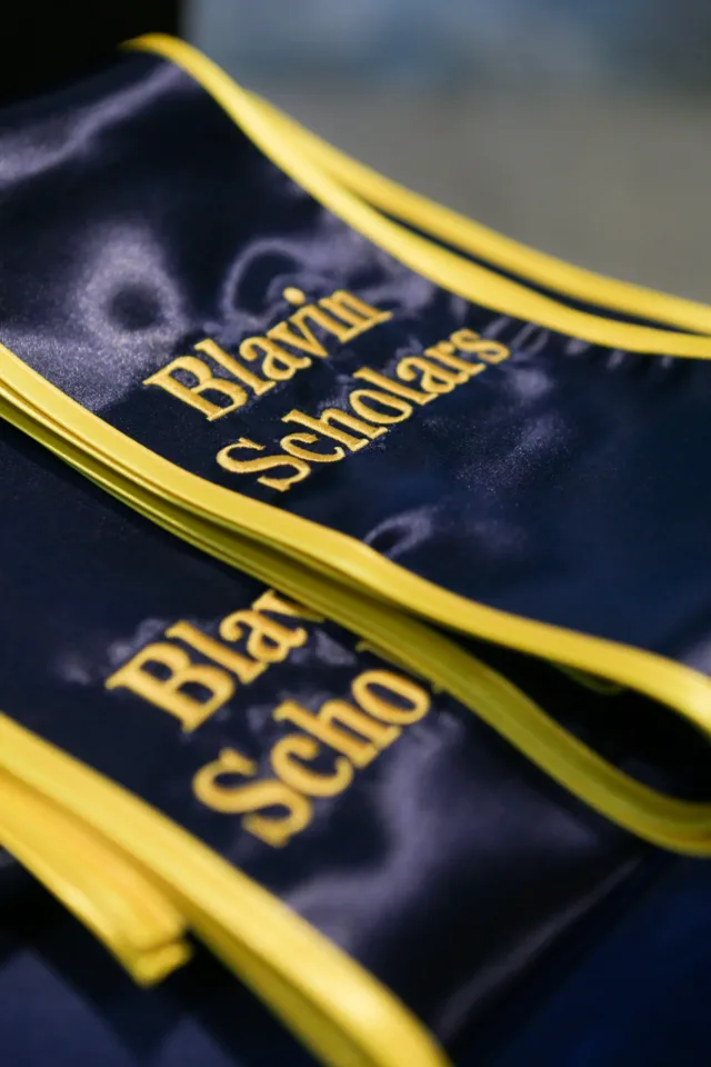 Close-up of folded blue graduation stoles with maize trim, featuring embroidered maize text that reads Blavin Scholars.