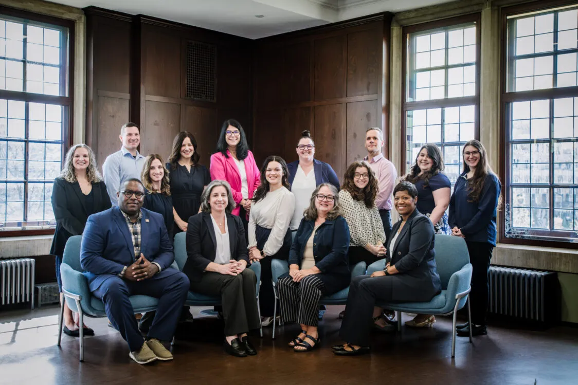 The Dean of Students staff pose for a photo. Some are seated in blue chairs while the rest stand against a backdrop of wood paneling. Sunlight filters in through the windows.
