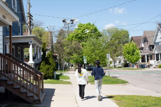 Two people walk down the sidewalk of a tree-lined street, passing various off-campus houses.