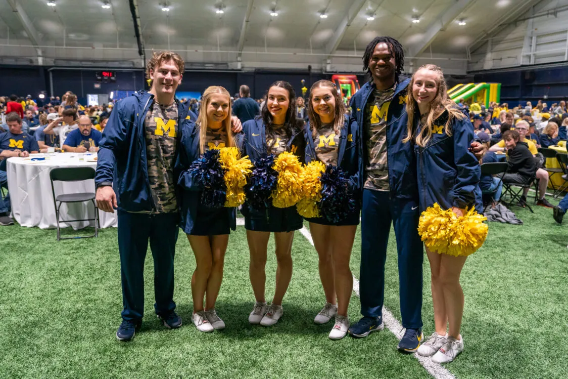 A group of six people, all dressed in Michigan gear, pose for a photo. Four of the people hold maize and blue pompoms.