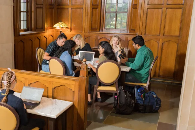 A small group of students sit in a wood-paneled study nook, gathered around a laptop and notebooks near a window.