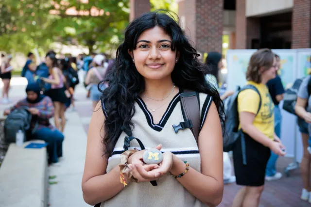 A student outdoors holds a small painted rock with a maize “M” in their cupped hands, with a backpack strap over one shoulder and other people blurred in the background.