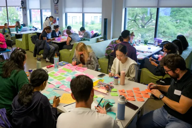 Students work together at tables covered in colorful sticky notes, writing and organizing ideas in a bright campus lounge with large windows.