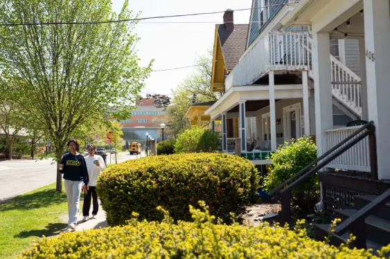 Two students holding coffee cups walk down a sunny sidewalk past residential student houses, with shoes hanging from overhead power lines and a yellow school bus visible in the background.