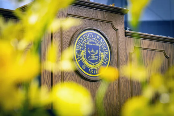 A close-up shot of the official University of Michigan seal mounted on a wooden podium, partially framed by softly blurred yellow flowers in the foreground.
