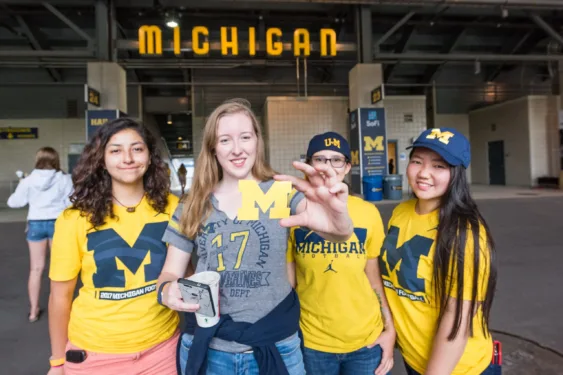 Four students, all dressed in maize and blue Michigan gear, stand in front of Michigan Stadium. One of the students holds a Block M cut out of paper.
