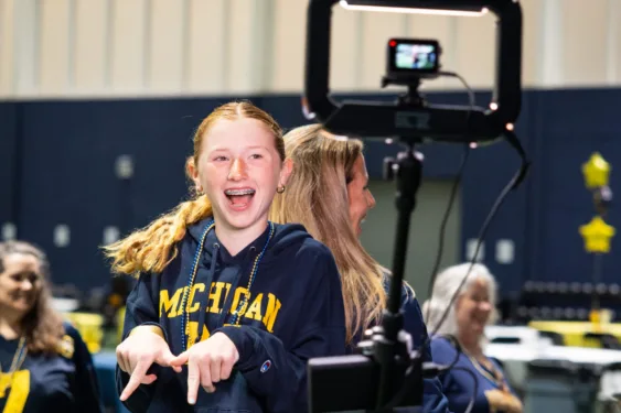 A student with light skin and long red hair is making an M with her hands. She is wearing a blue University of Michigan sweatshirt.