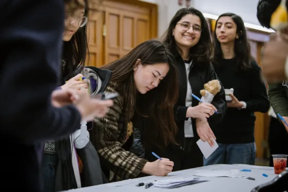 A student with long dark hair leans over a registration table to write on a piece of paper. Friends stand around waiting their turn to register.