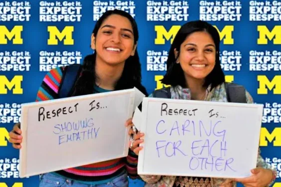 Two students, both with medium skin tone and longer black hair, smile for the camera. Behind them, a backdrop reads Give it. Get it. Expect Respect. The students are holding white boards. One reads Respect is showing empathy. The other reads Respect is caring for each other.