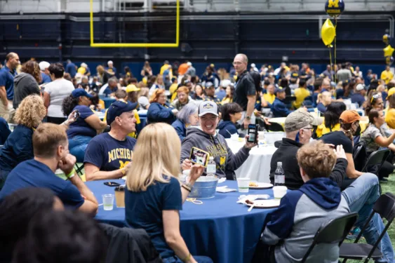 A large party is set up on an indoor field. Family members sit at tables and many of them are dressed in maize and blue Michigan wear. Two women smile at each other across a table in the foreground.