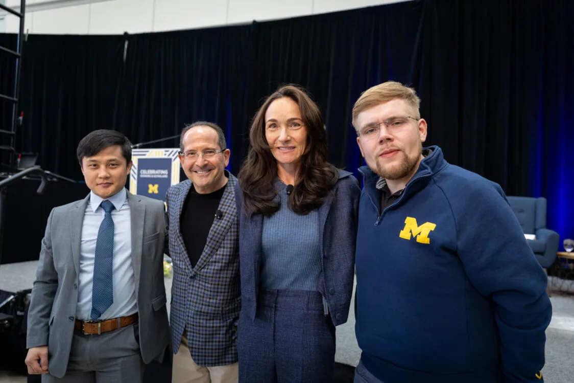 Paul and Amy Blavin pose with two Blavin Scholars. Paul is wearing a checkered sport coat and smiling broadly. Amy is wearing a blue sweater and a blue pantsuit. One scholar is wearing a suit and tie and has dark hair and skin. The other has light skin and is wearing a U-M sweatshirt.