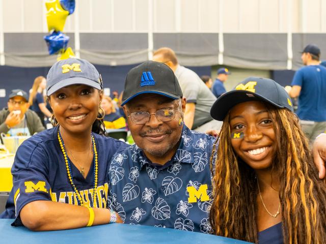 A family of three wearing University of Michigan gear smile while seated at a round table in the Oosterbaan Field House during the Parent & Family Weekend Tailgate